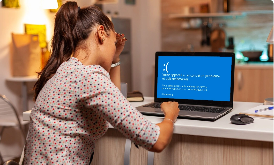 Une femme se tient la tête devant son ordinateur portable affichant un écran bleu d'erreur Windows